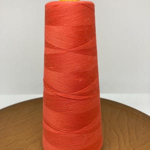 May include: A spool of bright orange thread, tightly wound around a cardboard core. The top of the spool is a bright yellow color. The thread is sitting on a wooden surface.