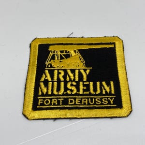 May include: A yellow and black embroidered patch with the text "Army Museum Fort DeRussy". The patch features a silhouette of a cannon.