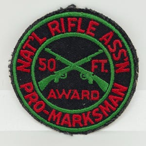 Puede incluir: Un parche redondo negro con texto e imágenes bordadas en rojo y verde. El texto dice "NATL RIFLE ASSN PRO-MARKSMAN AWARD 50 FT." Dos rifles verdes están cruzados en el centro.