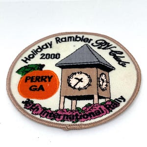 May include: Embroidered patch with a beige background and a brown border. The patch features a clock tower design with the text "Holiday Rambler RV Club 2000" and "6th International Rally". An orange fruit with the text "PERRY GA" is also present.