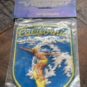 May include: Embroidered patch featuring a surfer riding a wave, with the word "California" in yellow script. The patch is blue with a yellow border and is packaged with a Voyager Originals header.