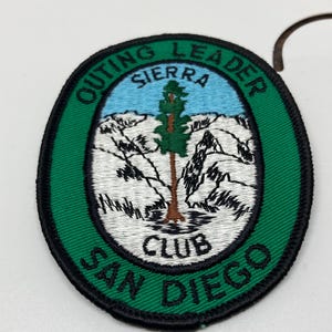 May include: Oval patch with a green border and black trim. The patch features the text "OUTING LEADER SIERRA CLUB SAN DIEGO". The centre design shows a tree, mountains, and a blue sky.