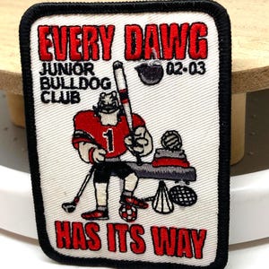 Puede incluir: Parche bordado con borde negro y texto "EVERY DAWG" en rojo. El parche presenta un bulldog de dibujos animados con uniforme rojo sosteniendo un bate de béisbol y un palo de golf. También incluye el texto "JUNIOR BULLDOG CLUB" y "HAS ITS WAY".