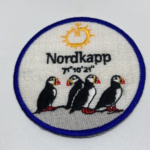 May include: Embroidered patch with a blue border, featuring four puffins. The patch has the word "Nordkapp" and coordinates above the puffins, with a gold sun design on a white background.