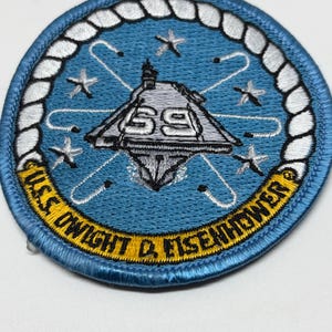 May include: A circular embroidered patch featuring a light blue background with a white rope border. The center displays a gray aircraft carrier with the number 69, surrounded by white stars and orbital lines. The text U.S.S. DWIGHT D. EISENHOWER is in yellow.