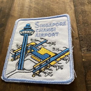 Pode incluir: Um remendo bordado branco com contorno azul. O remendo apresenta uma ilustração azul e amarela do Aeroporto de Changi de Singapura com o texto "Singapore Changi Airport".