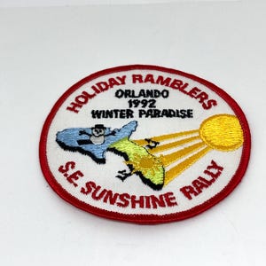 以下が含まれることがあります： 赤い縁取りの刺繍パッチ。「HOLIDAY RAMBLERS」と「SE SUNSHINE RALLY」の文字が特徴です。雪だるまと太陽のデザインの地図グラフィックと「ORLANDO 1992 WINTER PARADISE」の文字が含まれています。