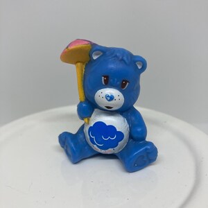 May include: A blue Care Bear figurine holding a yellow and pink umbrella. The bear has a white belly with a blue cloud design. The figurine is sitting on a white surface.
