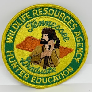 Può includere: Una toppa rotonda ricamata con un centro giallo e un bordo verde. La toppa presenta il testo "WILDLIFE RESOURCES AGENCY" e "HUNTER EDUCATION" in verde. Il centro raffigura una figura barbuta con un bastone.