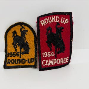 May include: Two vintage patches. One is yellow with black text and a silhouette of a person riding a horse, with the text "1956 ROUND-UP". The other is red with black text and a similar silhouette, with the text "ROUND UP 1954 CAMPOREE".