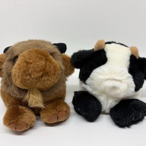 Puede incluir: Dos peluches redondos. Uno es un bisonte marrón de textura peluda, con orejas negras pequeñas y un mechón de pelo marrón claro en la barbilla. El otro es una vaca blanca y negra con cuernos pequeños de color tostado, orejas negras y una mancha.