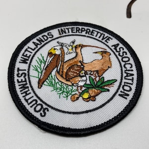 May include: Embroidered patch with a circular design featuring a brown pelican and other birds, green foliage, and the text "SOUTHWEST WETLANDS INTERPRETIVE ASSOCIATION" in black lettering on a white background, framed by a black border.