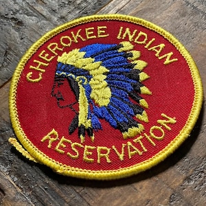 May include: A circular patch with the words "CHEROKEE INDIAN RESERVATION" embroidered in gold on a red background. The patch features a profile of a Native American in a headdress with blue and yellow accents.