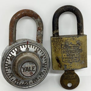 May include: A round silver-coloured Yale combination padlock with a rusted shackle next to a rectangular brass American Lock Company padlock with a dark shackle and a key inserted. The brass lock features an engraved shield logo and text.