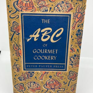 May include: Vintage cookbook titled "The ABC of Gourmet Cookery" by Peter Pauper Press. The cover is tan with orange and blue illustrations of food items. The title is in blue with white lettering.