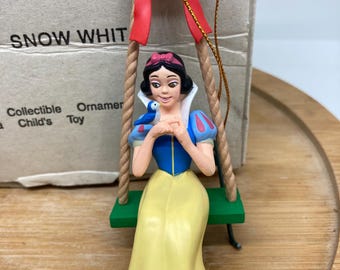 May include: Snow White Christmas ornament featuring a figurine of Snow White sitting on a swing with a blue bird. She is wearing a yellow dress with a blue bodice and a red bow in her hair. The ornament is made of plastic and has a gold string for hanging.