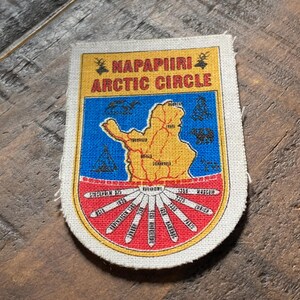 Op de afbeelding: Rechthoekige patch met de tekst "NAPAPIIRI ARCTIC CIRCLE". De patch heeft een gele rand, een blauw midden met een kaart en een rood onderste gedeelte met stralende lijnen en tekst. De patch ligt op een houten oppervlak.