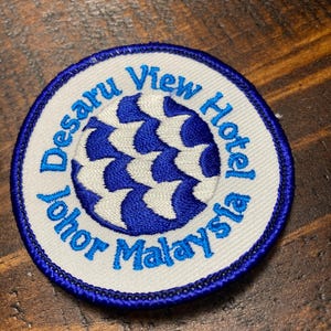 Puede incluir: Un parche bordado blanco con un borde azul. El parche presenta un diseño de remolino azul y blanco en el centro y el texto "Desaru View Hotel Johor Malaysia" alrededor del borde.