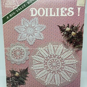 Op de afbeelding: Een vintage boekcover met de titel "DOILIES!" toont witte gehaakte kleedjes in verschillende ontwerpen tegen een roze bloemenachtergrond. De tekst van de American School of Needlework is ook zichtbaar.