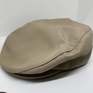 May include: A tan-colored, flat cap made of a smooth fabric. The cap has a rounded crown and a small, stiff brim. The hat is a classic style, often worn for a casual or semi-formal look. The cap is a solid color with no visible patterns or embellishments.