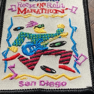 May include: Embroidered patch featuring a running musician playing a guitar, with the text "SUZUKI Rock 'N' Roll Marathon San Diego." The patch has a black border and a cream-colored background with colorful accents.