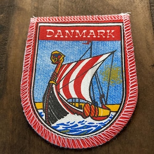 May include: A red, white, and blue embroidered patch with the word "DANMARK" in white lettering. The patch features a Viking ship with a red and white striped sail sailing on blue water.