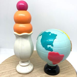 May include: A colorful toy ice cream cone with a pink ball on top of orange spheres, set on a white base. Beside it is a small globe with continents labeled in teal and red, on a black base.