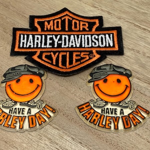 May include: A Harley-Davidson patch and two smiley face pins. The patch is black and orange with the words "MOTOR HARLEY-DAVIDSON CYCLES". The pins have orange smiley faces wearing hats, with the text "HAVE A HARLEY DAY!"