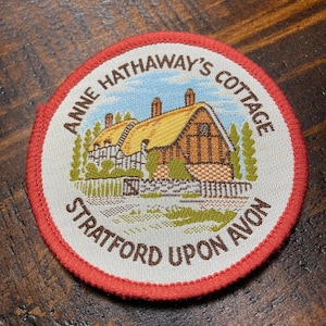 May include: A white and red circular patch with a brown embroidered image of Anne Hathaway's Cottage in Stratford-upon-Avon. The text "Anne Hathaway's Cottage" is embroidered in brown around the top of the patch and "Stratford-upon-Avon" is embroidered in brown around the bottom of the patch.