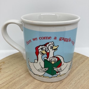 May include: White ceramic mug with a light blue background featuring three cartoon geese wearing Santa hats and holding a green book. The mug has the text "Here we come a gaggle-ing" in black. The mug has a white handle and rim.