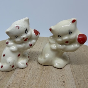 May include: A pair of off-white ceramic cat figurines. The cat on the left features red polka dots and a red accent on its back. The cat on the right holds a solid red ball. Both cats have painted black eyes and red mouths, with a glossy finish.