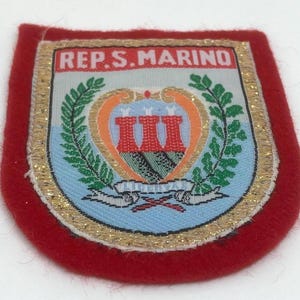 May include: Red felt patch with a gold embroidered border. The patch features a crest with a white background and a red and orange shield with three towers. The text "REP. S. MARINO" is above the crest.