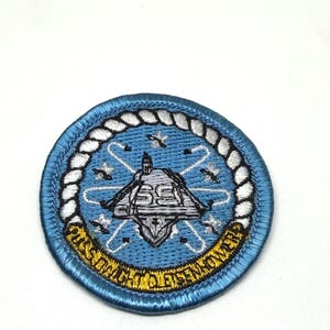 May include: A circular embroidered patch featuring a light blue background with a white rope border. The center displays a gray ship silhouette with the number 69, surrounded by white atomic symbols. A yellow banner at the bottom reads USS DWIGHT D EISENHOWER.
