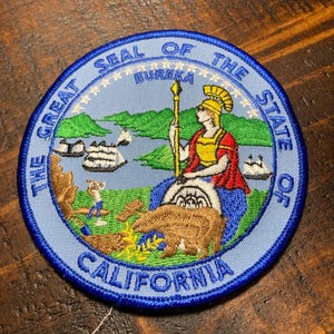 May include: A blue and white embroidered patch with the Great Seal of the State of California. The seal features a woman in a red and gold robe holding a spear, a bear, and a landscape with ships and mountains. The text "The Great Seal of the State of California" and "Eureka" are embroidered on the patch.