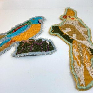 Puede incluir: Parches bordados de pájaros. Un parche presenta un pájaro azul, naranja y verde sobre una base verde. El otro parche representa un pájaro marrón, amarillo y verde posado en una rama verde. Ambos parches tienen un borde gris.