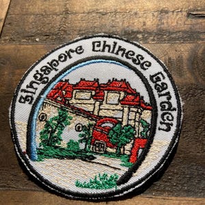 Pode incluir: Um remendo bordado branco com contorno preto que representa um edifício vermelho com um céu azul e árvores verdes. O texto "Singapore Chinese Garden" está escrito em preto ao redor da borda do remendo.