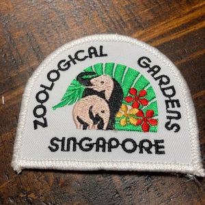 Może przedstawiać: Biała naszywka haftowana z tekstem "Zoological Gardens Singapore" w czarnych literach. Naszywka przedstawia wzór zielonego i białego liścia palmy z dwoma różowymi słoniami i czerwonymi kwiatami.