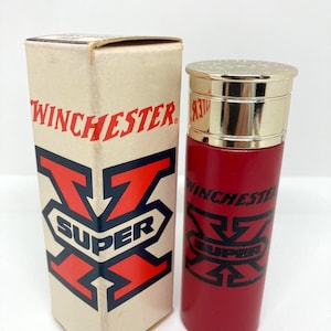 Puede incluir: Una botella roja Winchester Super con tapa dorada y caja a juego. La caja y la botella presentan el logotipo de Winchester en negro y rojo. La botella es cilíndrica y la caja es rectangular.