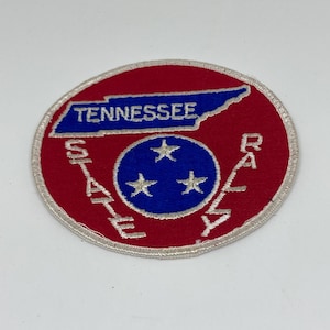 Op de afbeelding: Een cirkelvormige patch met een rode achtergrond en witte stiksels. De patch heeft een blauwe omtrek van Tennessee met het woord "TENNESSEE" erboven. Binnen de cirkel bevindt zich een blauwe cirkel met drie witte sterren en de woorden "STATE RALLY".