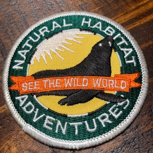 Puede incluir: Un parche bordado verde y blanco con fondo amarillo. El parche presenta una foca negra con un sol blanco detrás. El texto en el parche dice "Natural Habitat Adventures See the Wild World".