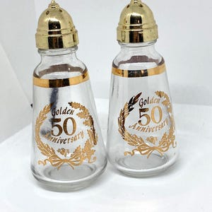 Puede incluir: Un par de saleros y pimenteros de vidrio transparente con tapas de metal dorado. Cada salero tiene una banda dorada alrededor de la parte superior y presenta el texto "Golden 50 Anniversary" en escritura dorada y un diseño de corona dorada.