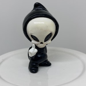Pode incluir: Uma pequena figura preta de um personagem semelhante à Morte, com um rosto de caveira branco e um capuz preto. A figura está a fazer um gesto obsceno com uma mão pintada de branco. Sobre uma superfície redonda branca.