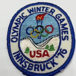 May include: A circular patch with a blue border and white background. The patch features the text "OLYMPIC WINTER GAMES INNSBRUCK '76 USA". The center design includes a torch, Olympic rings, and a mountain scene.