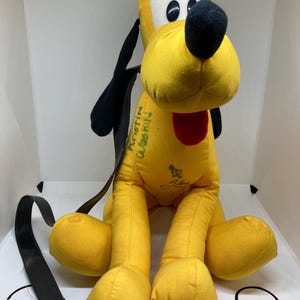 May include: A plush toy of Pluto, a yellow dog character with black ears and a black nose. The toy has a white face, black eyes, and a red tongue. It has a black strap and green text on the side.