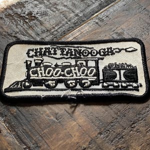 May include: A rectangular patch with a black border and a white background. The patch features a black illustration of a train with the words "CHATTANOOGA" above it and "CHOO-CHOO" below. The patch is likely for a jacket or bag.