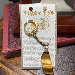Op de afbeelding: Een gouden sleutelhanger met een hanger van bruin en gouden tijgeroog. De steen heeft een chatoyance-effect, met een gouden glans. De kaart die aan de sleutelhanger is bevestigd, vermeldt "Tiger Eye" en bevat informatie over de oorsprong en eigenschappen van de steen.