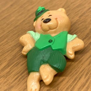 Könnte beinhalten: Kleine, hellbraune Bärenfigur mit grüner Weste, Shorts und Hut mit Kleeblatt. Der Bär hat einen lächelnden Ausdruck und ist mit ausgestreckten Armen posiert. Die Figur besteht aus glänzendem Material.