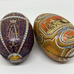 May include: Two oval-shaped decorative tins. The left tin is dark blue with gold filigree patterns and a prominent letter A topped with a crown. The right tin is orange and gold, featuring detailed illustrations of classical figures and scenes.