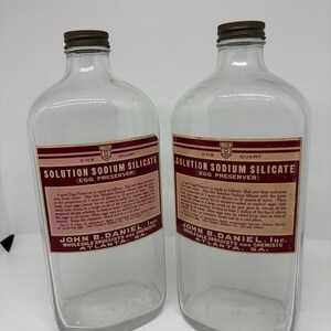 May include: Two clear glass bottles with brown metal caps, each featuring a rectangular label with red and white text reading Solution Sodium Silicate Egg Preserver, John B. Daniel, Inc. Wholesale Druggists and Chemists Atlanta, GA, one quart.