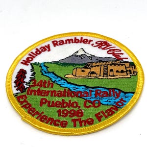 May include: Embroidered patch with a yellow border featuring text and a landscape scene. The patch reads "Holiday Rambler RV Club" and "34th International Rally Pueblo, CO 1996 Experience The Flavor."